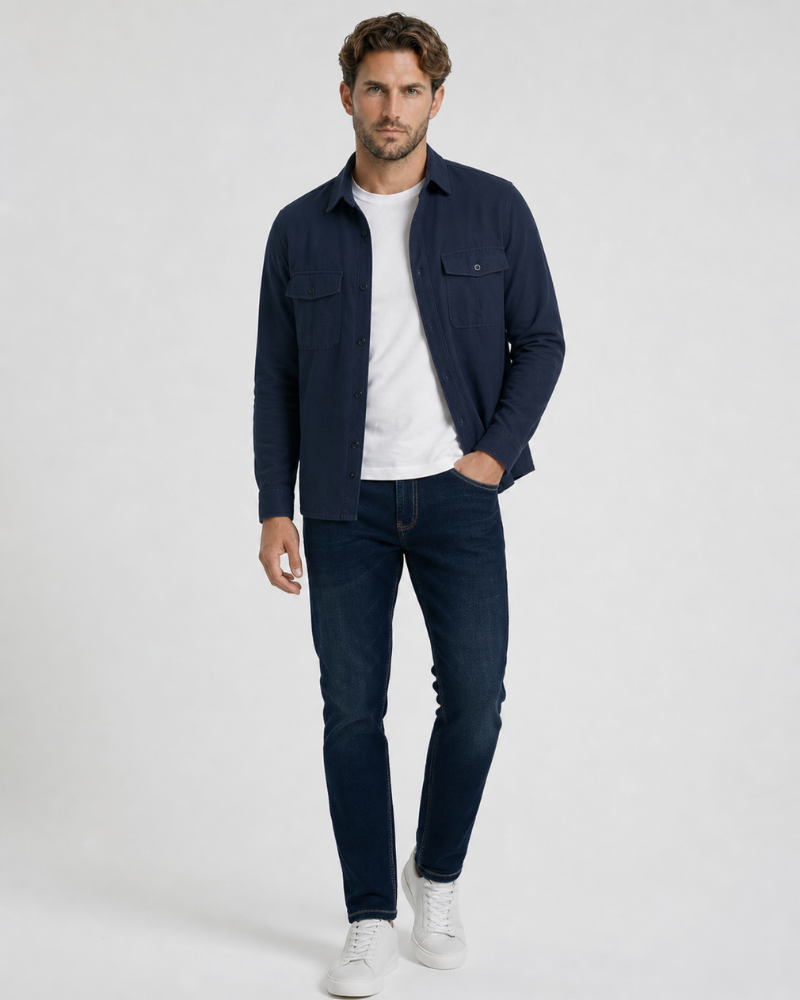Man wearing a By Alden navy shirt over a white t-shirt, dark jeans, and white sneakers on a light gray background #color_navy