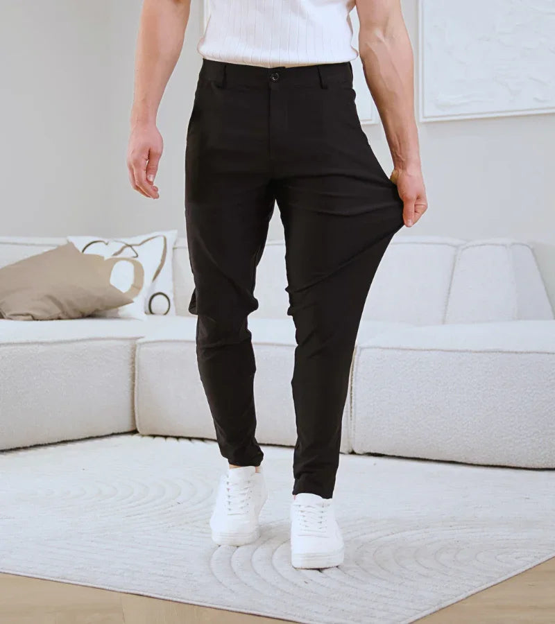 Man wearing black slim fit pants and white sneakers pulling pant leg to show stretch in modern living room