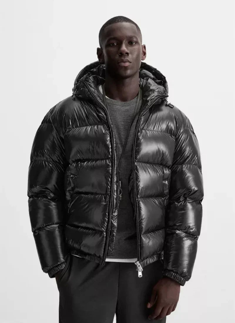 Male model wearing Alden Ash black shiny down puffer jacket with hood, gray sweater, and black pants