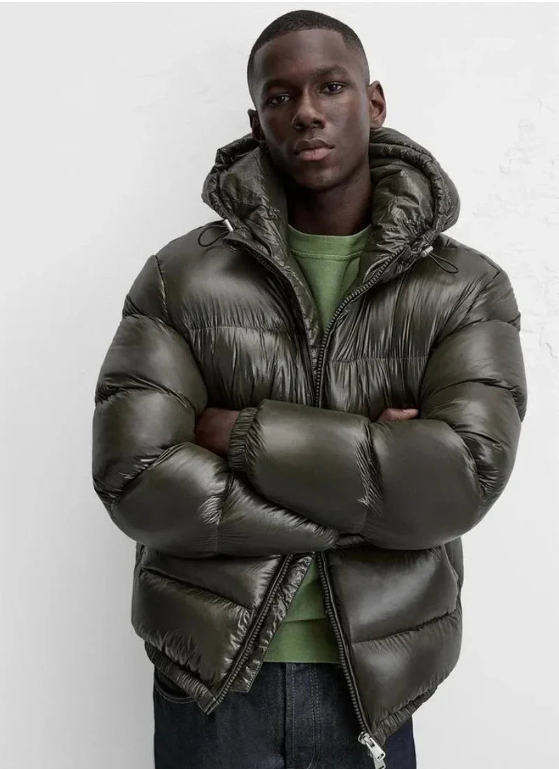 Man wearing shiny dark olive puffer jacket with hood and green sweatshirt, standing against white wall