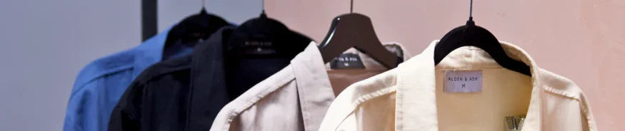 Four collared shirts in blue, black, cream, and beige on black hangers against a pink wall