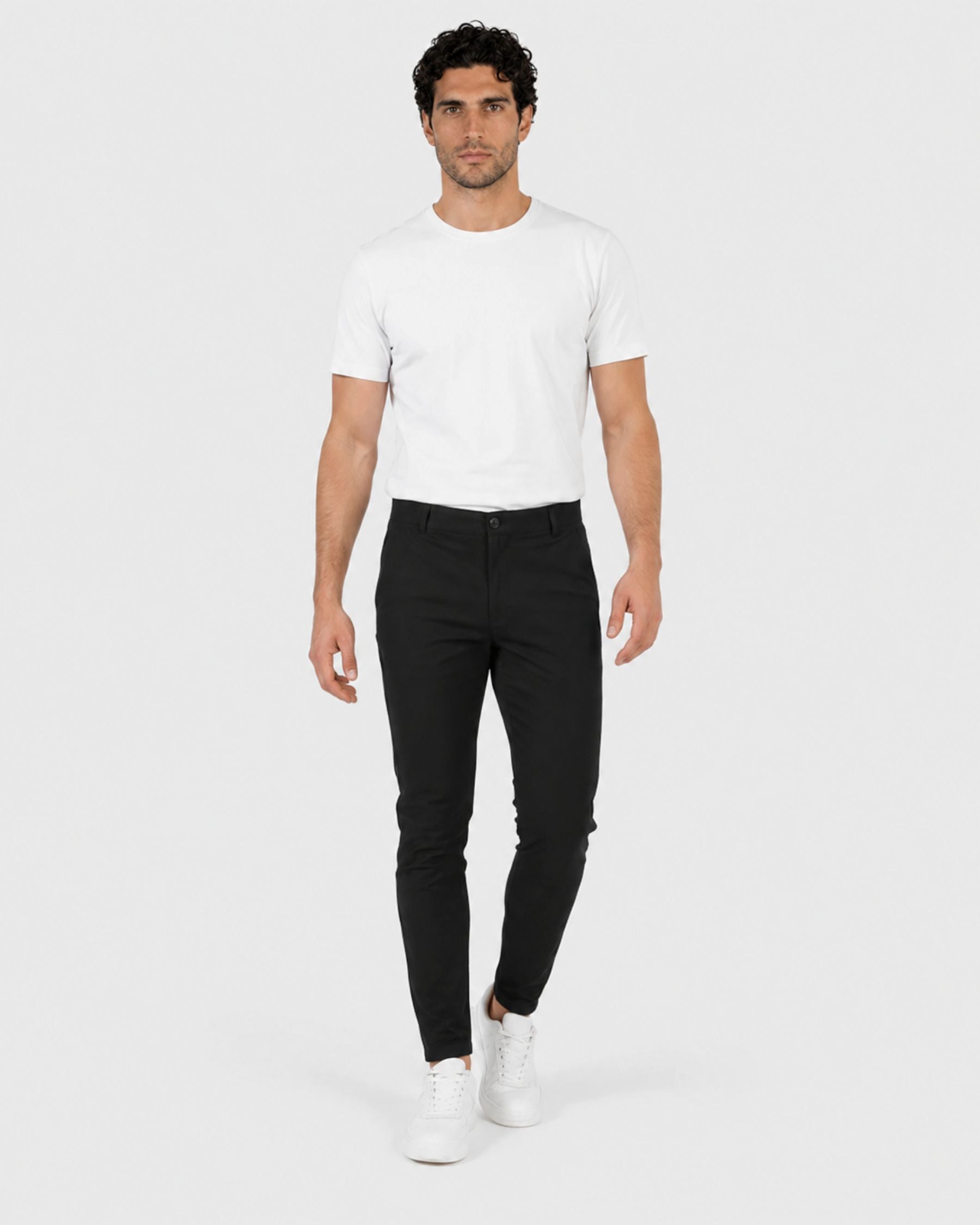 A man modeling a white crew-neck t-shirt and slim black trousers with white sneakers, standing against a light gray studio background.