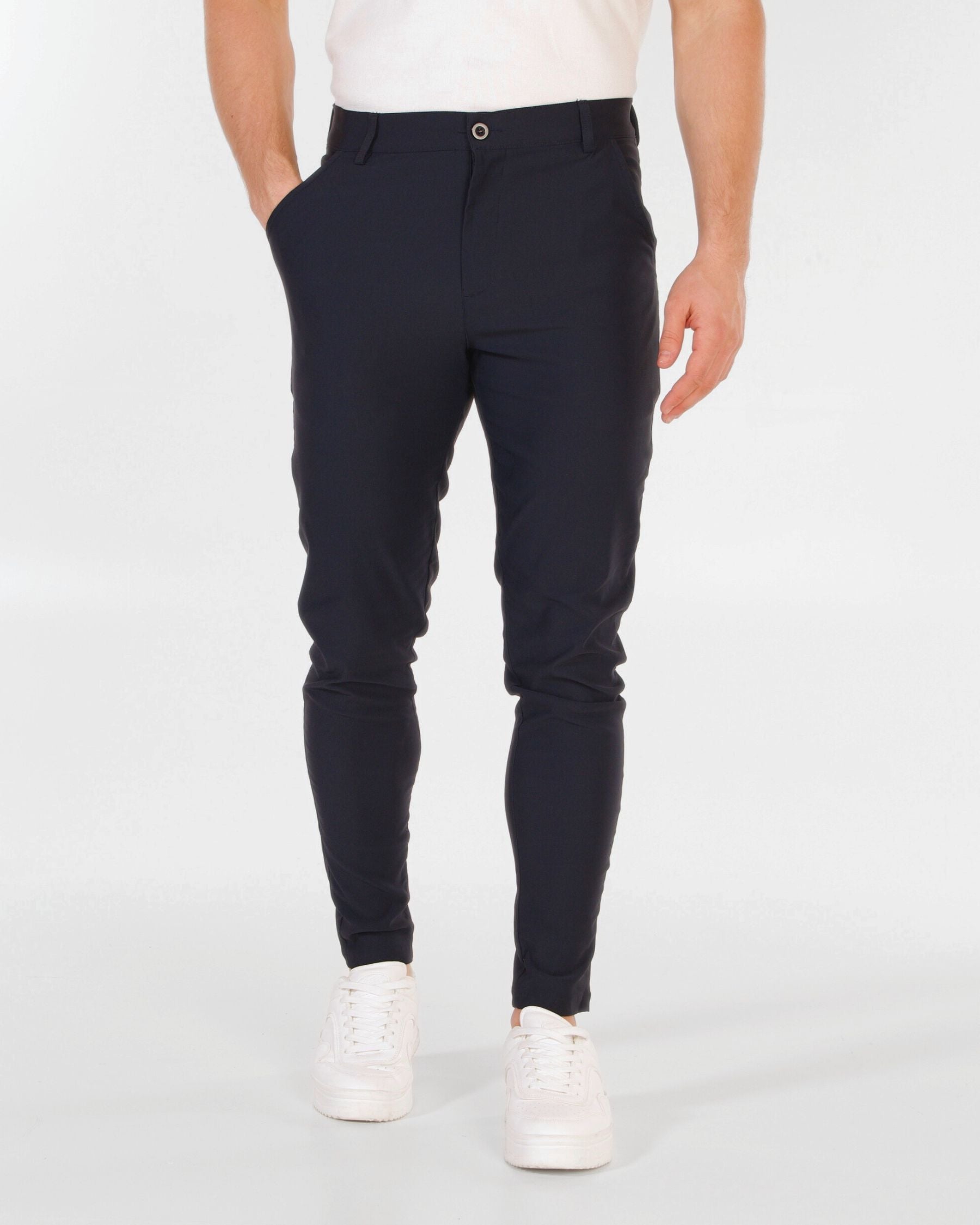 A person wearing slim-fit dark navy trousers with a white t-shirt and white sneakers, shown from the waist down against a light gray studio background.