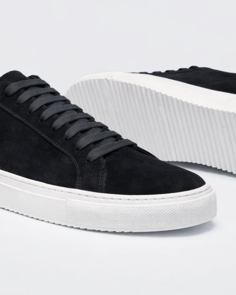 Close-up of minimalist luxury black suede trainers highlighting the textured white rubber sole and premium stitching.