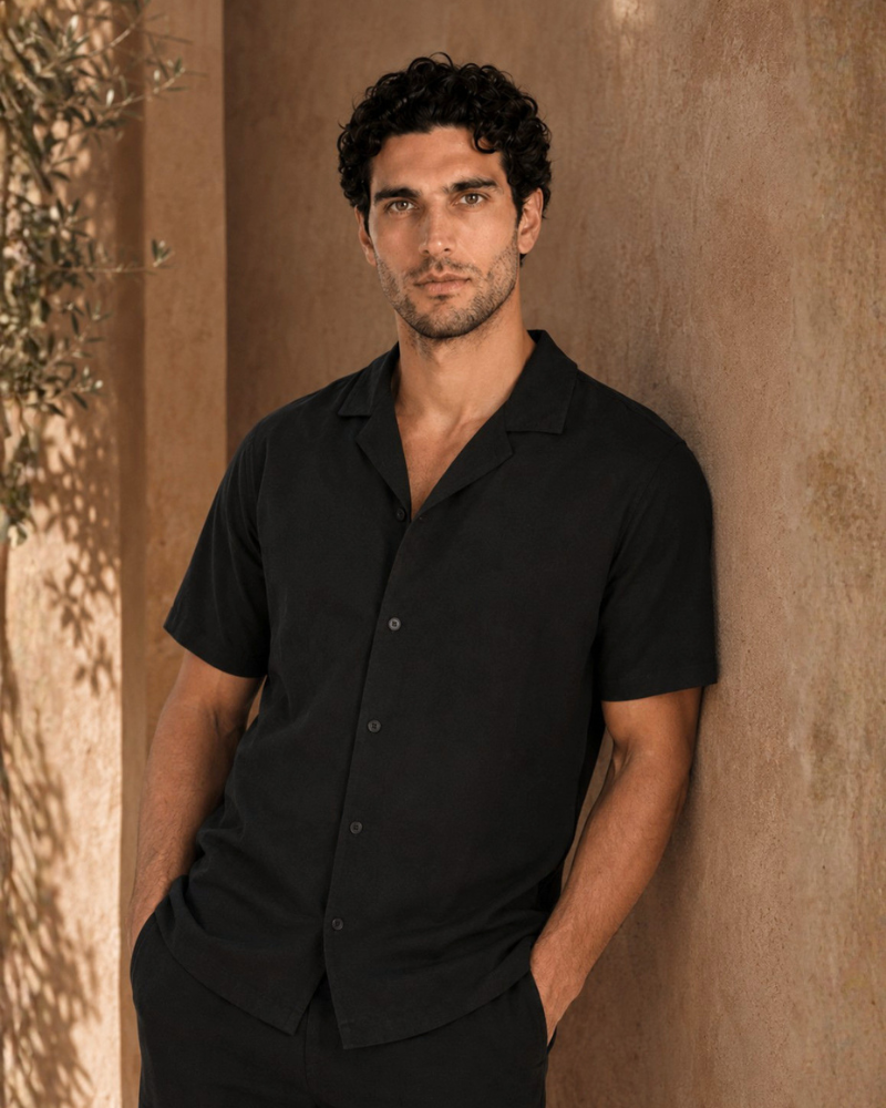 By Alden black short-sleeve camp collar shirt on male model, front view, upper body, warm beige wall background.
#Color_black