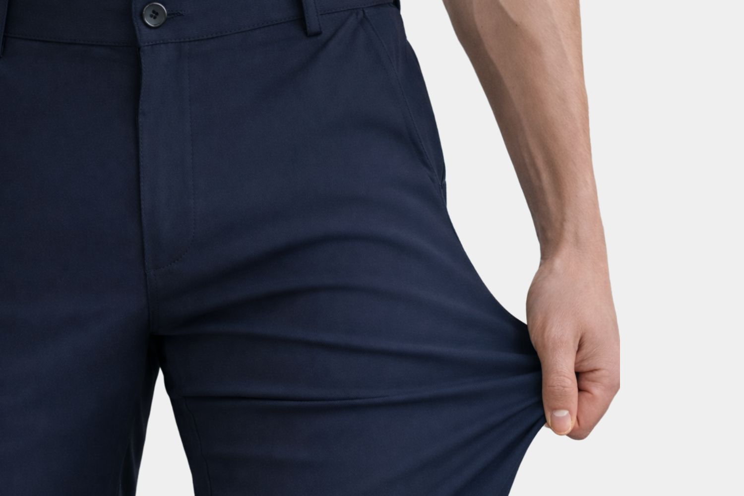 Close-up of a person wearing dark navy trousers, pulling the fabric at the thigh to demonstrate stretch, against a light gray background.